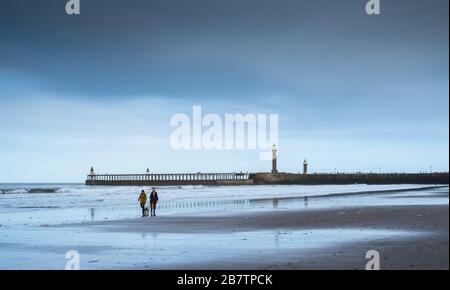 Due persone camminano i loro cani sulla spiaggia di Whitby, con il molo di Whitby sullo sfondo, costa dello Yorkshire settentrionale, Inghilterra, Regno Unito Foto Stock