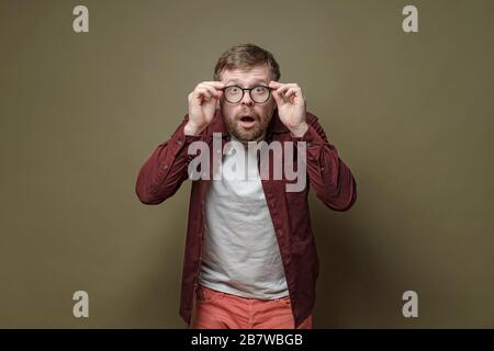 L'uomo bearded sorpreso, in una camicia di borgogna, in stupore tiene gli occhiali nelle mani e guarda con gli occhi grandi nella macchina fotografica. Foto Stock