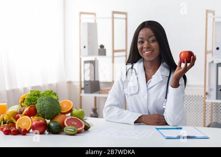 African american sorridente dottore dietologo seduto al banco di lavoro, tenendo la mela Foto Stock