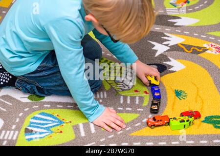 Ragazzo di cinque anni che gioca e allineano le auto giocattolo su un tappetino da gioco con le strade. Il ragazzo è vestito di jeans blu e tiene una macchina gialla. Foto Stock