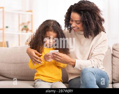 Madre Africana che insegnano la figlia per usare lo smartphone seduto sul divano Foto Stock