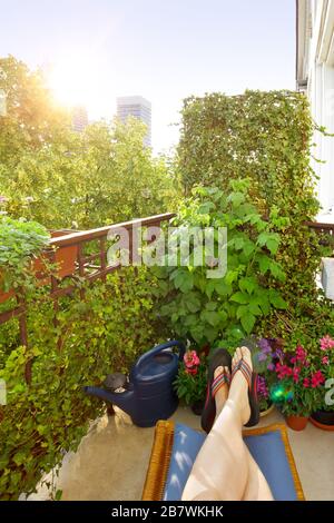 Balcone della città in estate: Piedi della donna in infradito su un poggiapiedi e un sacco di piante e fiori in pentole. Foto Stock