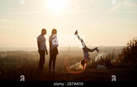 Madre e padre sono in piedi sulla collina e guardando la loro figlia facendo un cavalletto, dietro di loro bella vista orizzonte di un tramonto del sole Foto Stock