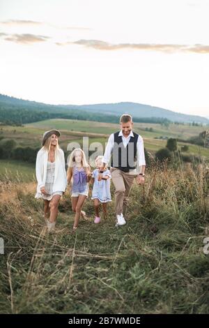 Famiglia felice in abiti casual, madre, padre e due piccole figlie sulla natura, camminando insieme tenendo le mani, su mer tramonto in campo selvaggio in Foto Stock