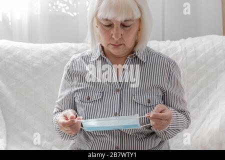 Donna bionda adulta in una camicia a righe mette una maschera medica sul suo viso in una stanza a casa.Virus protezione, coronavirus, ARVI concetto Foto Stock