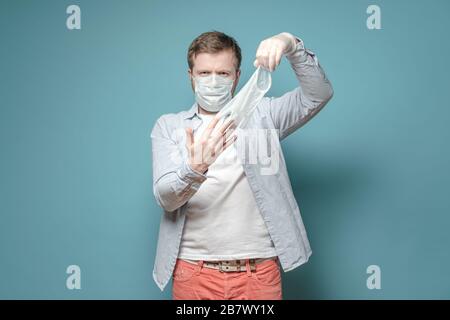 Uomo serio in una maschera medica, toglie i guanti di lattice usati, su uno sfondo blu. Concetto di sicurezza e protezione dai virus. Foto Stock