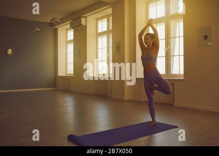 Yoga donna pratica yoga in piedi su una gamba medita l'equilibrio in una stanza con finestre soleggiate. Foto Stock