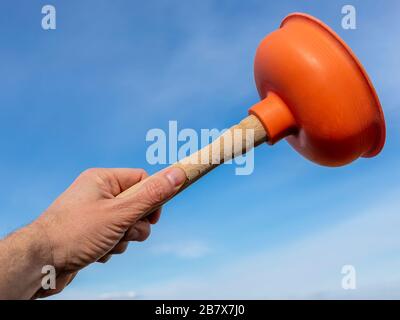 La mano di un uomo bianco solleva uno stantuffo arancione verso il cielo blu, tenendolo per il manico di legno Foto Stock