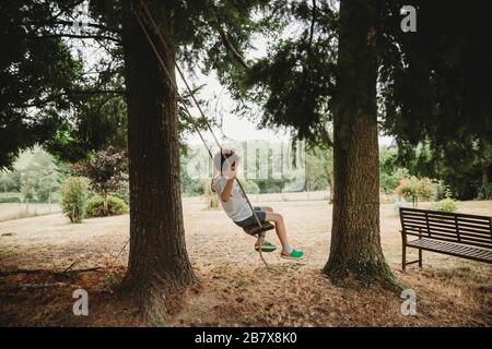 Vista laterale del ragazzo su swing sotto gli alberi Foto Stock