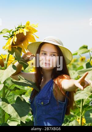 Ragazza adolescente in abito denim con sorriso mostra sul girasole. Campo agricolo. Scelta. Verticale Foto Stock