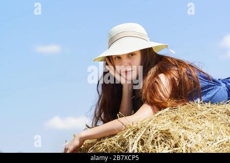 Una bella ragazza adolescente in un ampio cappello da sposa e vestito blu si trova su un rotolo di paglia nel campo. Guarda la fotocamera. Primo piano Foto Stock