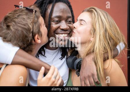 Due ragazze bionde baciano un ragazzo africano da un muro. Gruppo multiculturale Foto Stock