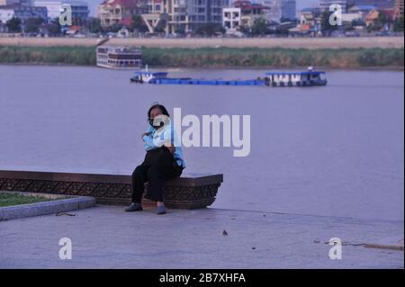 Una donna cambogiana anziana in maschera si siede da sola sul fiume deserto durante la pandemia di coronavirus. Phnom Penh, Cambogia. © Kraig Lieb Foto Stock