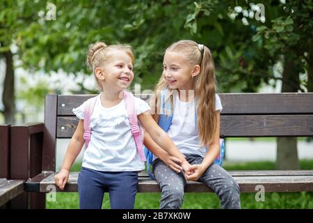 Due bambini ridono e chiacchierano su una panchina. Ragazze con zaini. Il concetto è di ritorno a scuola, famiglia, amicizia e infanzia. Foto Stock