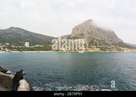 Un uomo fotografa la vista dal Golitsyn Trail al villaggio turistico di Novy Svet nella Crimea. Giorno nuvoloso all'inizio della primavera Foto Stock