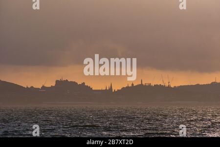 Firth of Forth, Scozia, Regno Unito. 18 marzo 2020. Regno Unito Meteo: Un colorato orange al tramonto sopra il caratteristico profilo delle guglie di Edimburgo vista attraverso il quarto estuario da East Lothian Foto Stock