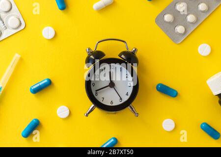Sveglia, pillole bianche, capsule blu, ago della siringa e flacone su sfondo giallo. Epidemia, antidolorifici, concetto sanitario. Iniezione medica. Foto Stock