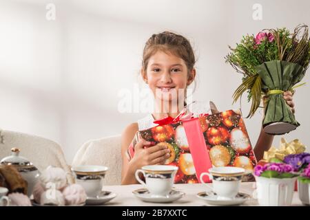 Bella ragazza positiva che tiene un regalo di Capodanno nelle sue mani e un bouquet di fiori seduti al tavolo delle feste durante il tè bevendo. Anno nuovo Foto Stock