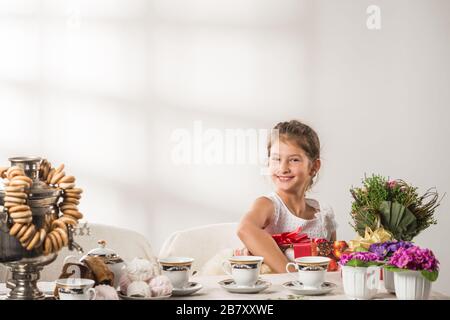 Bella ragazza positiva che tiene un regalo di Capodanno nelle sue mani e un bouquet di fiori seduti al tavolo delle feste durante il tè bevendo. Anno nuovo Foto Stock