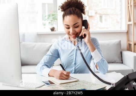 Agente di tour femminile di discesa africana seduto al tavolo in ufficio che parla con il cliente sul telefono fisso prendendo appunti durante il viaggio sorridendo allegro Foto Stock