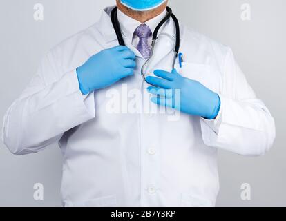 medico maschile adulto in un cappotto medico bianco si alza e tiene uno stetoscopio nero su uno sfondo bianco, spazio copia Foto Stock