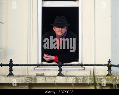 Uomo in un cappotto e cappello trilby guarda fuori dalla sua finestra mentre fuma Foto Stock