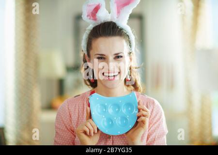 Ritratto di giovane donna sorridente in una blusa rosa e spighe conigliate di pasqua a casa moderna nel giorno di primavera soleggiato mostrando piatto a forma di pollo blu per le uova. Foto Stock