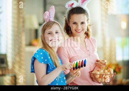 sorridente giovane madre e figlia in orecchie di coniglietto pasquale con cesto con uova da fattoria e cibo colorazione istruzioni di lettura nel moderno soggiorno in s Foto Stock