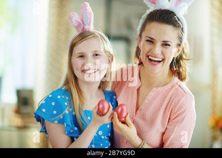 Ritratto di felice madre elegante e figlia in spighe conigliate di pasqua nella casa moderna in sole primavera giorno mostrando le uova rosse di pasqua. Foto Stock