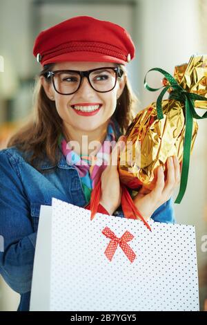 donna trendy e pensiva di 40 anni in una camicia jeans e cappello rosso in occhiali con avvolto in foglio d'oro grande uovo di pasqua e bianco polka puntino shopping bag in Foto Stock