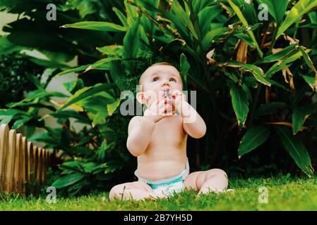 Baby nel pannolino senza vestiti seduto sull'erba rodendo un fiore. Foto Stock