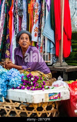 Un venditore si inclina oltre al suo stallo nella produzione di mercato in Ubud, Bali. Lei è circondata dal colore dei fiori e il materiale Foto Stock