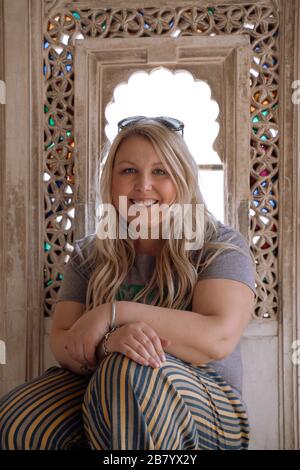 Felice, donna sorridente con lunghi capelli biondi siede di fronte ad una finestra di vetro colorato ornato. Presa in Udaipur India Foto Stock