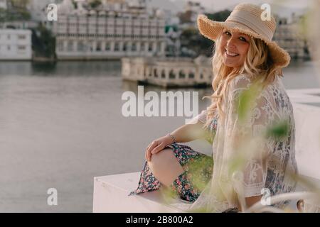 Donna bionda sorridente che indossa una parte superiore di merletto con un cappello di paglia pone che domina il lago di Pichola in Udaipur India Foto Stock