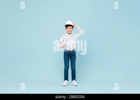Ritratto a tutta lunghezza di sorridente giovane asiatico ragazzo aspirando ad essere futuro ingegnere indossando bianco hardHat tenendo blueprint in blu isolato studio backgro Foto Stock