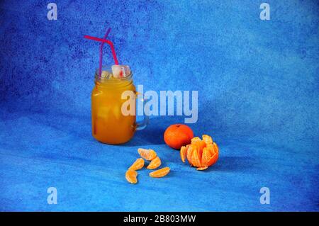 Una tazza di succo di mandarino con ghiaccio e cannucce accanto a pelati e tutta la frutta e fette di mandarino su uno sfondo blu. Close-up. Foto Stock