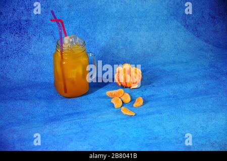 Una tazza di succo di mandarino con ghiaccio e cannucce accanto a pelati e tutta la frutta e fette di mandarino su uno sfondo blu. Close-up. Foto Stock