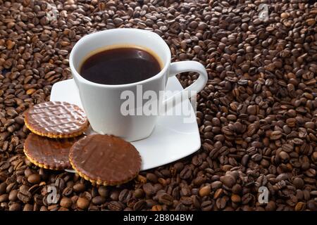 Tazza bianca di caffè caldo su uno sfondo di fagioli marroni. Tre biscotti al cioccolato. Foto Stock