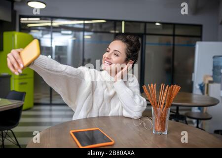Giovane donna seduta al tavolo da cucina, facendo selfie Foto Stock