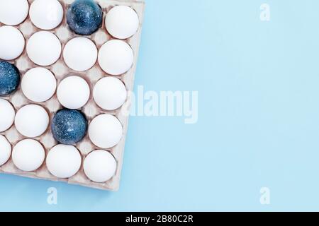 Molte uova bianche e alcune dipinte in blu si trovano nel contenitore su sfondo azzurro. Vista dall'alto. Concetto di Pasqua. Spazio di copia. Foto Stock