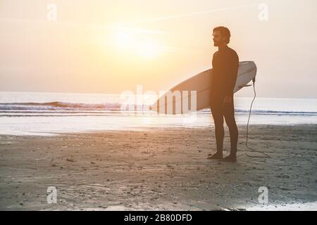 Silhouette di surfista in piedi sulla spiaggia in attesa di onde al tramonto - uomo con tavola da surf indossando muta wet che guarda l'alba - Extreme sport conc Foto Stock
