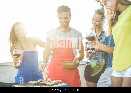 Felice studenti che cucinano e bevono vino al barbecue sul tetto cena - migliori amici grigliare carne con salsa barbecue - gioventù, cibo, amicizia concetto - Fo Foto Stock