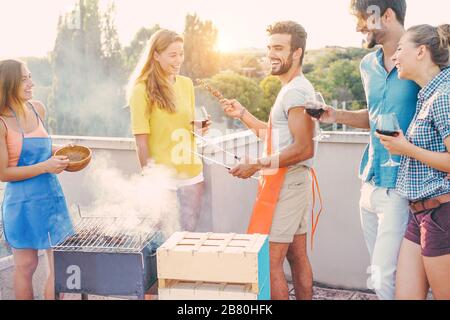 Amici felici che cucinano la carne del barbecue e bevono il vino sulla parte superiore del tetto alla festa del barbecue - i giovani che preparano la cena all'aperto - fuoco su due GU del centro Foto Stock