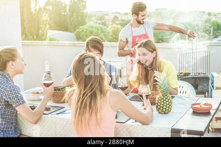 I giovani amici mangiano e bevono vino rosso al barbecue sul tetto - gente felice grigliare e divertirsi al barbecue cena all'aperto - principale attenzione sulla righ Foto Stock