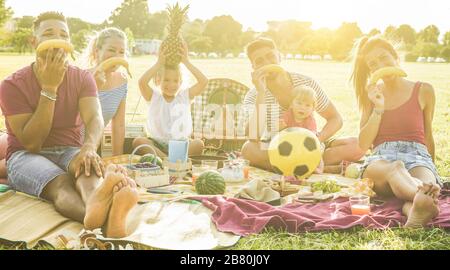 Amici di famiglia felici che giocano con la frutta mentre fanno i volti divertenti al picnic all'aperto di fine settimana - i genitori che si divertono con i loro bambini che mangiano e ridono Foto Stock