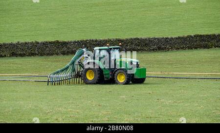 Coltivatore impegnato a lavorare in azienda agricola, guida trattore verde spandendo slurry con tubi flessibili trailing per fertilizzare terreno agricolo pascolo campo - Yorkshire, Inghilterra, Regno Unito. Foto Stock