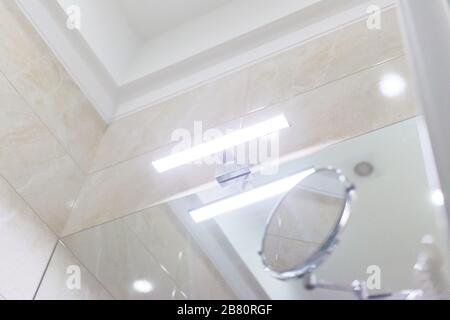 Interni del bagno, dettaglio primo piano. Parte del soffitto a cassettoni, luce a LED sopra lo specchio del lavandino, specchio cosmetico per il trucco, piastrelle in marmo beige su Th Foto Stock