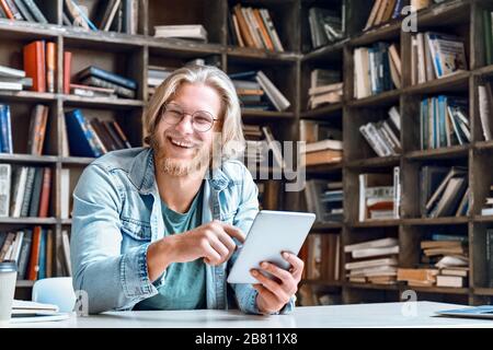 Felice studente di sesso maschile studiare online risata tenere tablet digitale in biblioteca, ritratto. Foto Stock