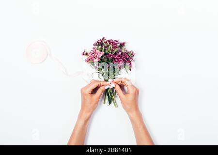 Mano donna che lega nastro satinato fiocco su piccolo bouquet su scrivania bianca, vista dall'alto. Foto Stock