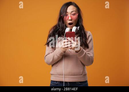 Brunetta asiatica in occhiali rosa con telefono in mano isolato in studio su sfondo arancione vuoto Foto Stock
